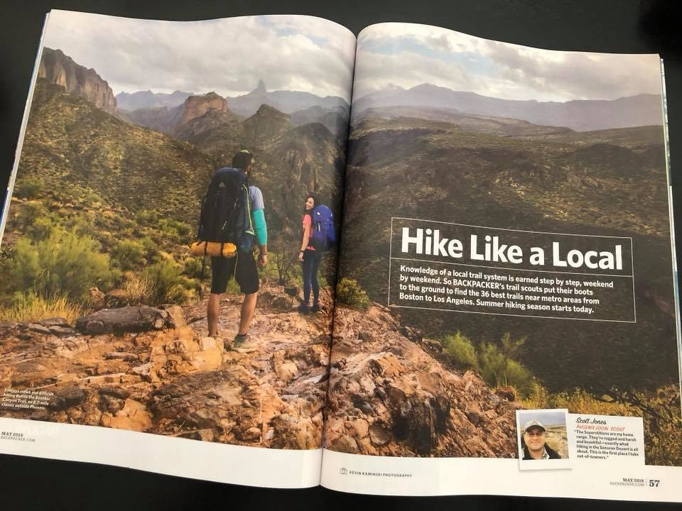 Auto-generated description: Two hikers walk along a rocky trail in a vast, mountainous landscape under a cloudy sky.