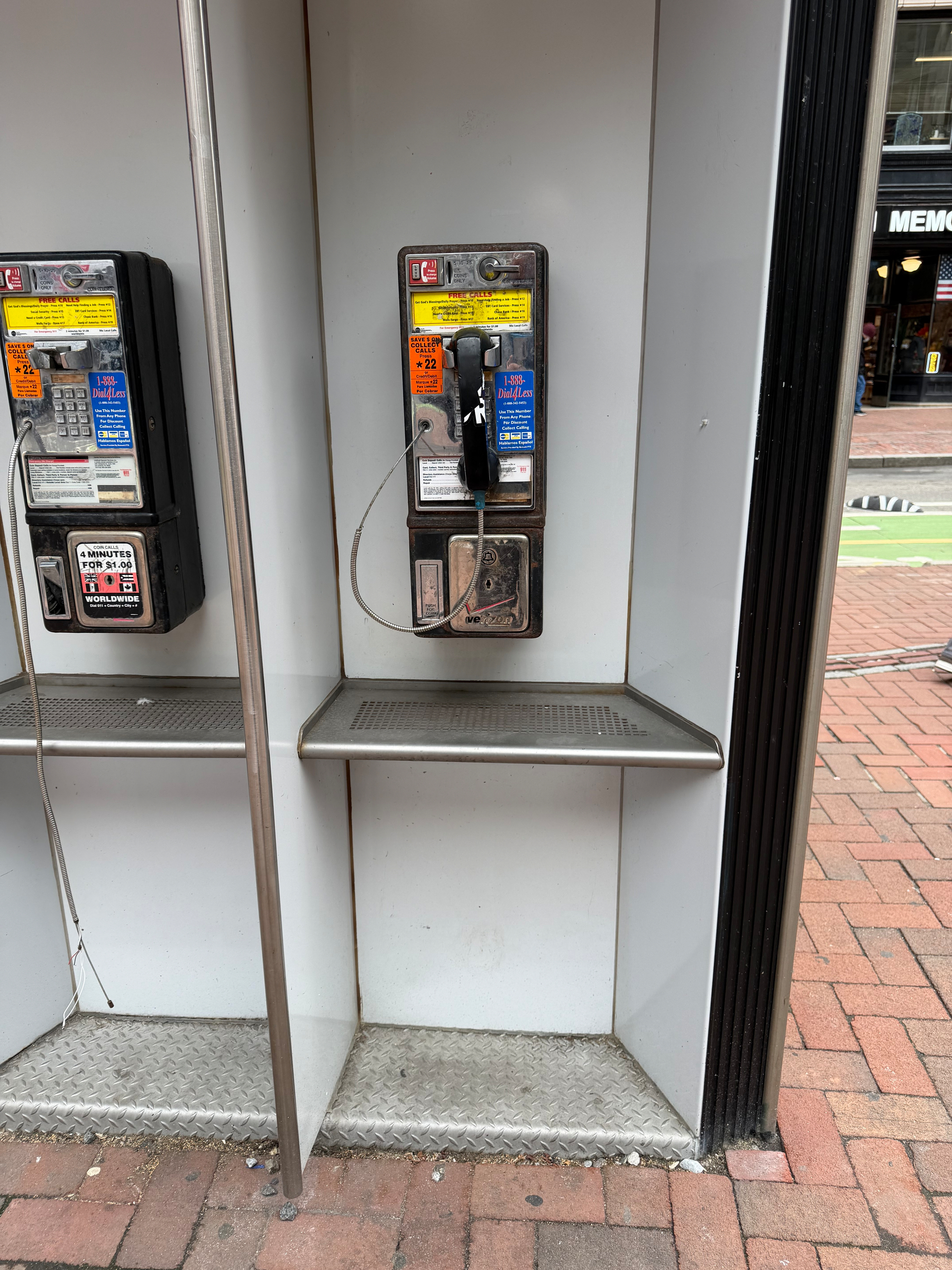 Two public payphones are mounted side by side in an outdoor booth with a brick pavement in front.