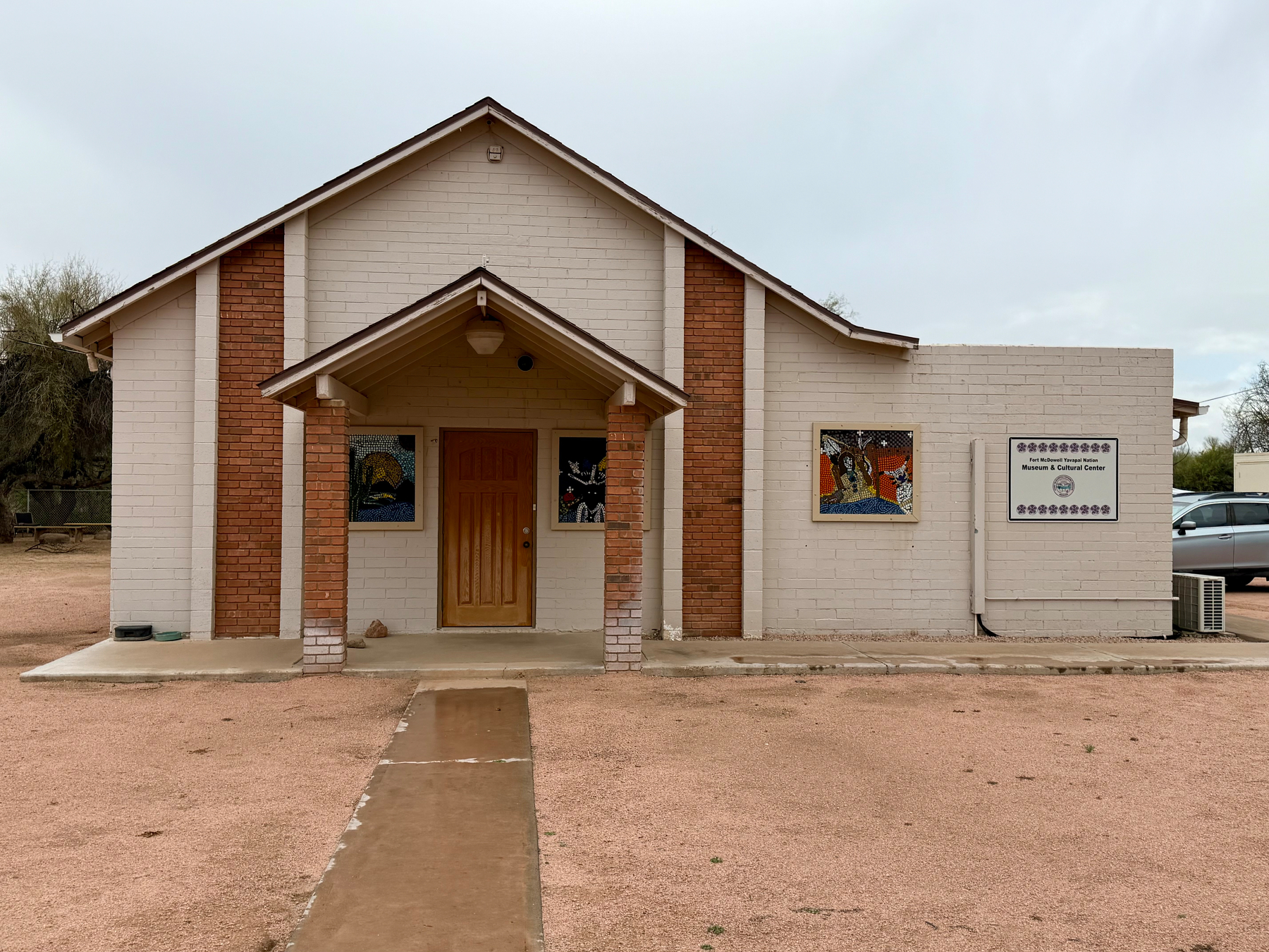 Auto-generated description: A small, single-story brick building with an entrance canopy and two colorful murals on the front facade stands in a desert-like environment.