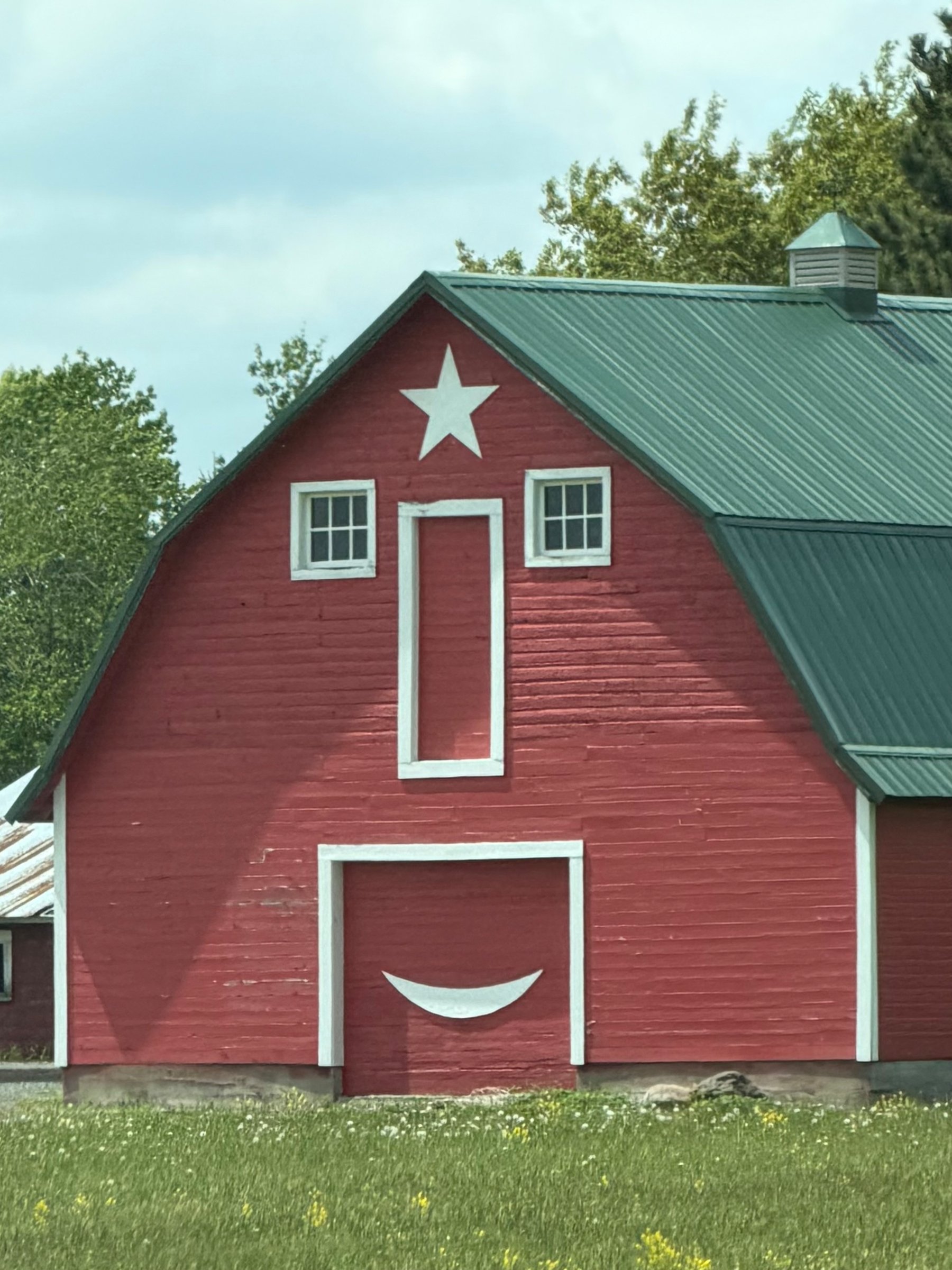 A red barn features a face-like design with two square windows as eyes, an elongated window as a nose, and a smiley shape as a mouth, topped with a star.