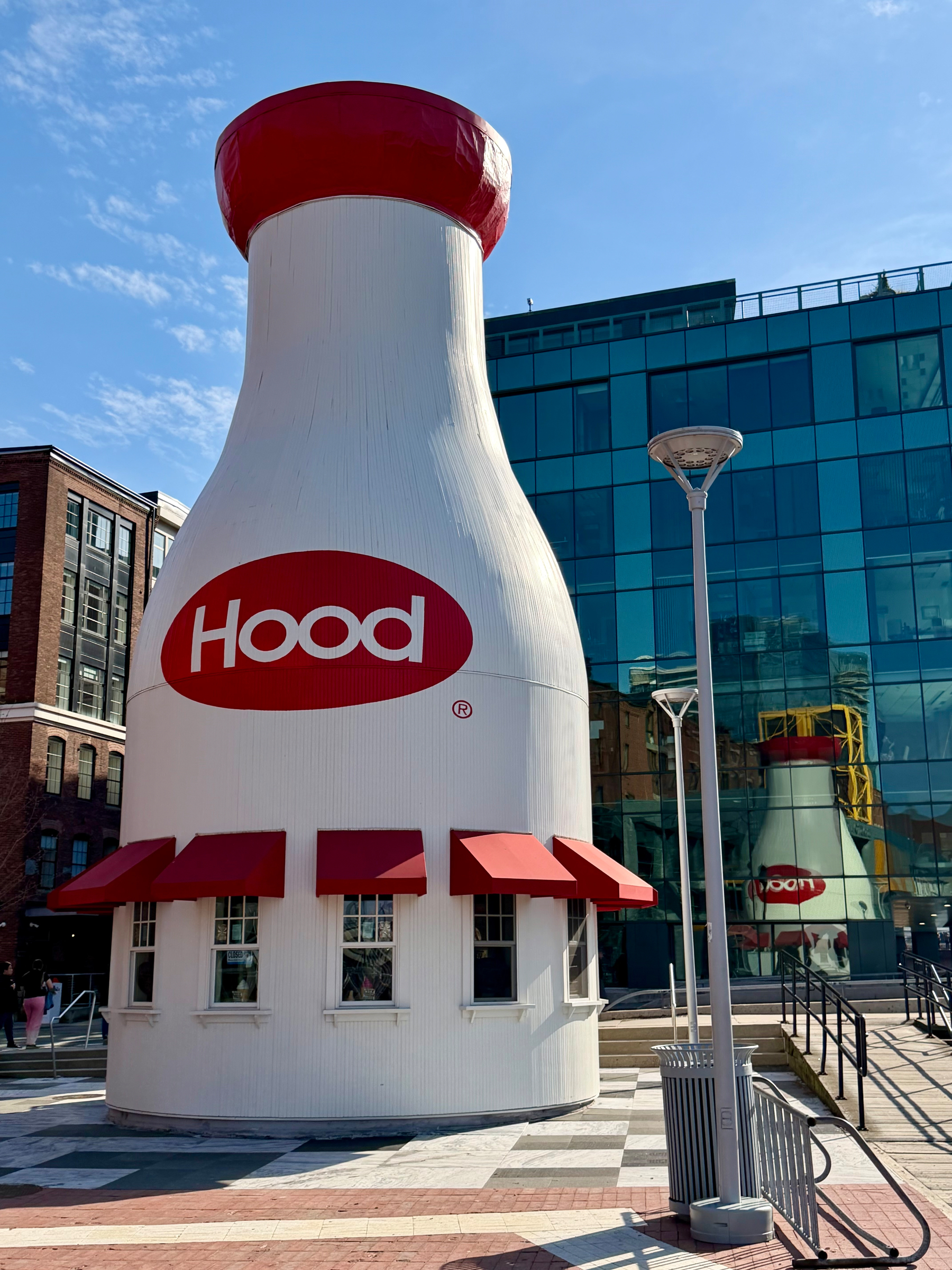 A large, building-sized structure resembling a milk bottle with the Hood logo is situated in an urban area with modern architecture and streetlights.