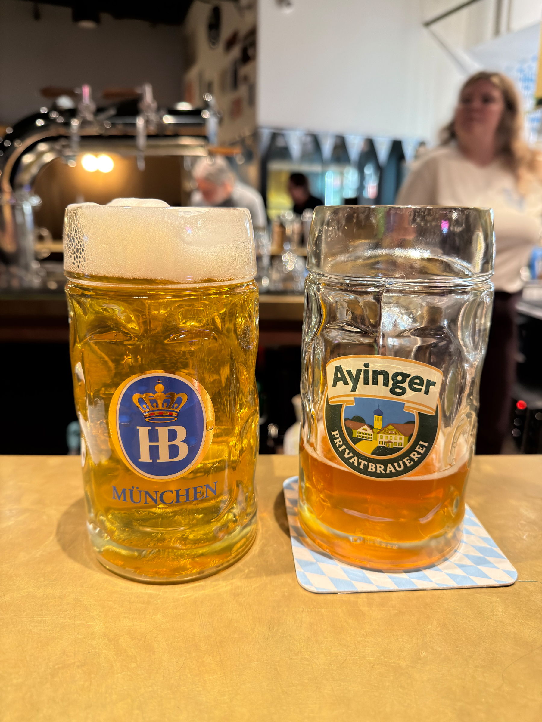 Two large beer mugs filled with different types of beer are placed on a bar counter, one labeled HB München and the other Ayinger Privatbrauerei.