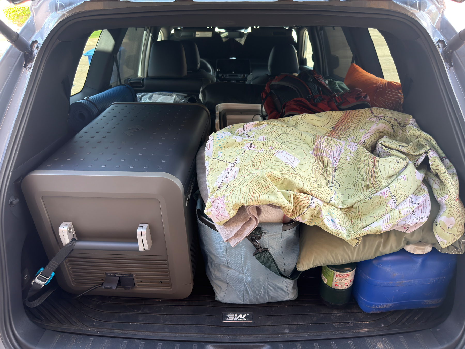 The back of an SUV is packed with various camping or travel gear, including a cooler, bags, blankets, and containers.