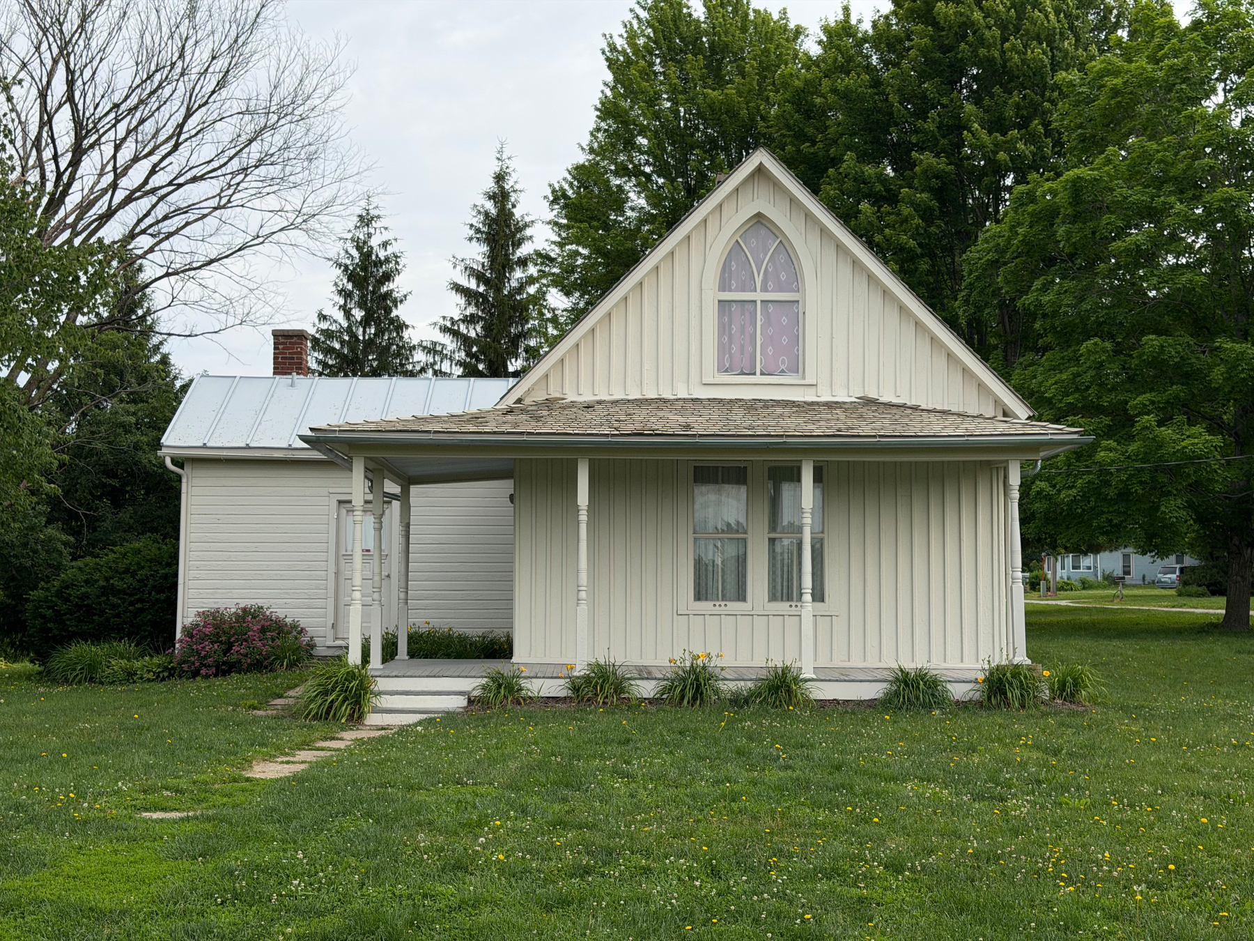A quaint, small house with a steep Gothic-style gable roof and surrounding green lawn is set amidst trees and a cloudy sky. This is the inspiration for the background of the American Gothic painting.