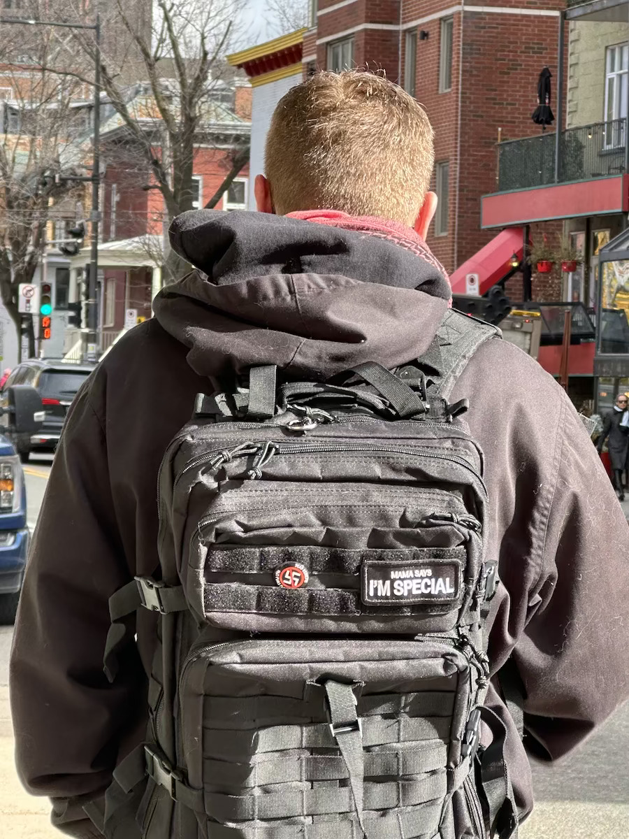 A young man carrying a black backpack with a tiny anti-fascist button attached