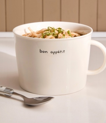 A white mug with bon appétit inscribed on it contains a serving of noodles topped with herbs, accompanied by a spoon.