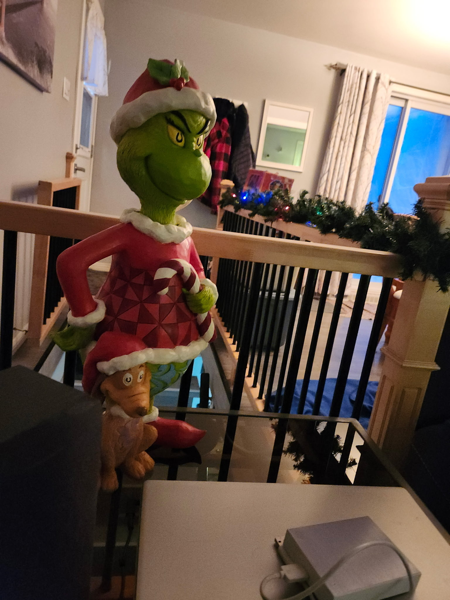 A Grinch statue dressed in a Santa outfit stands on a staircase railing beside a decorated living room with greenery.
