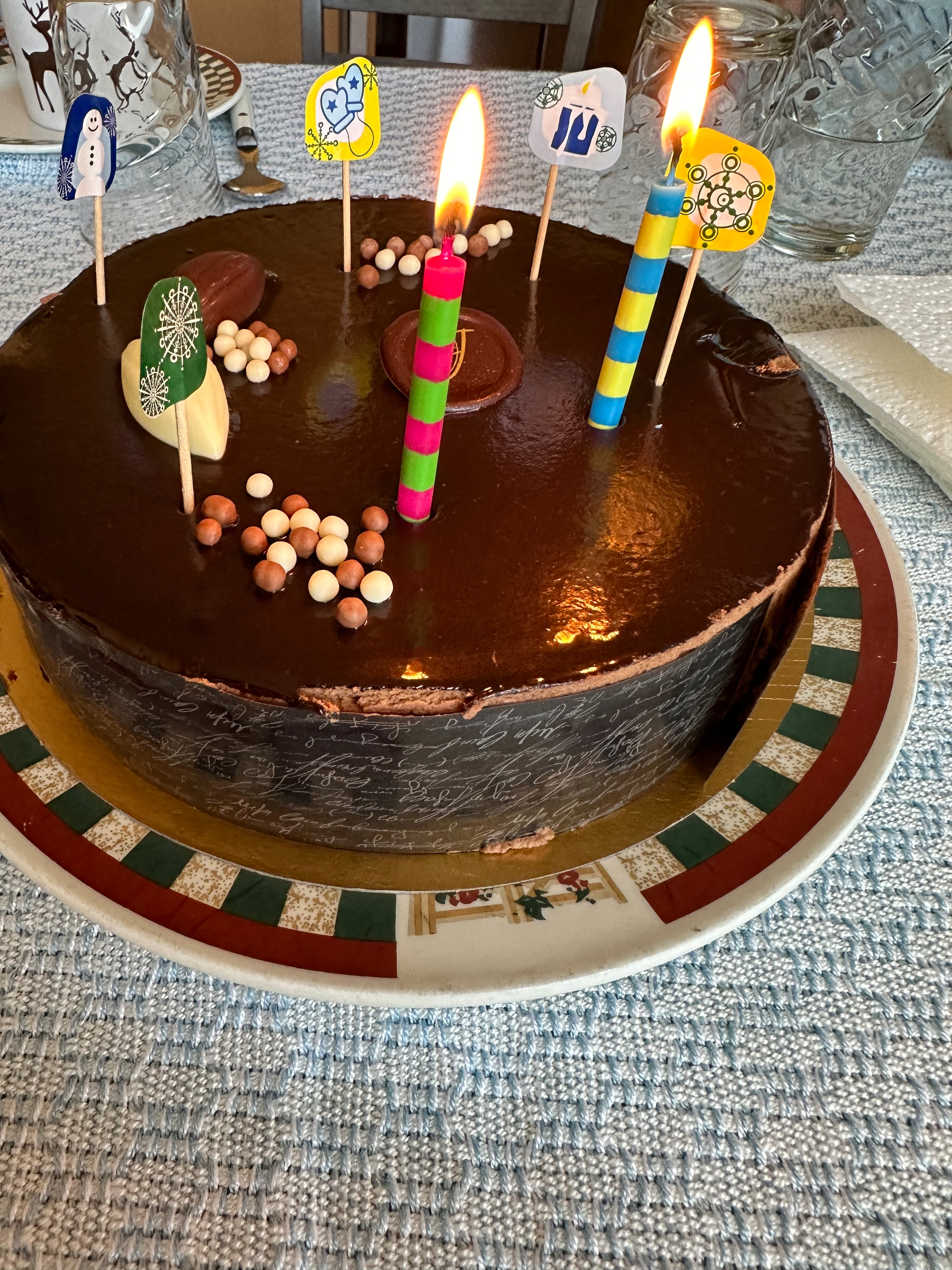 A chocolate cake with three lit candles and decorative toppers is placed on a colorful plate.