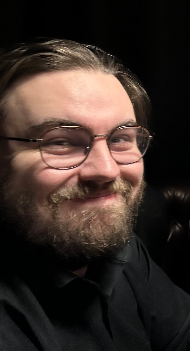A person with glasses and a beard is smiling, seated against a dark background.