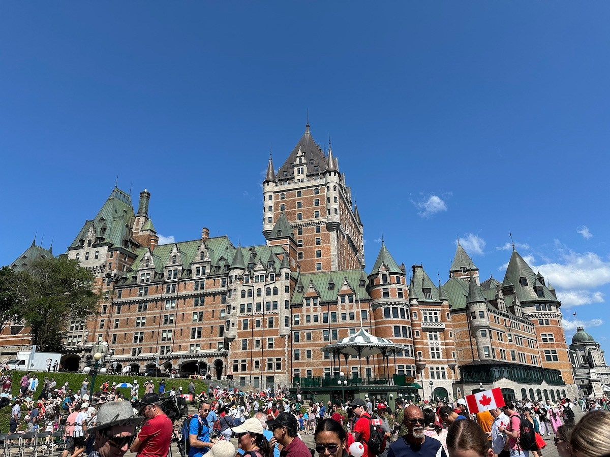 A large crowd gathers outside the historic Château Frontenac under a clear blue sky in Quebec City.