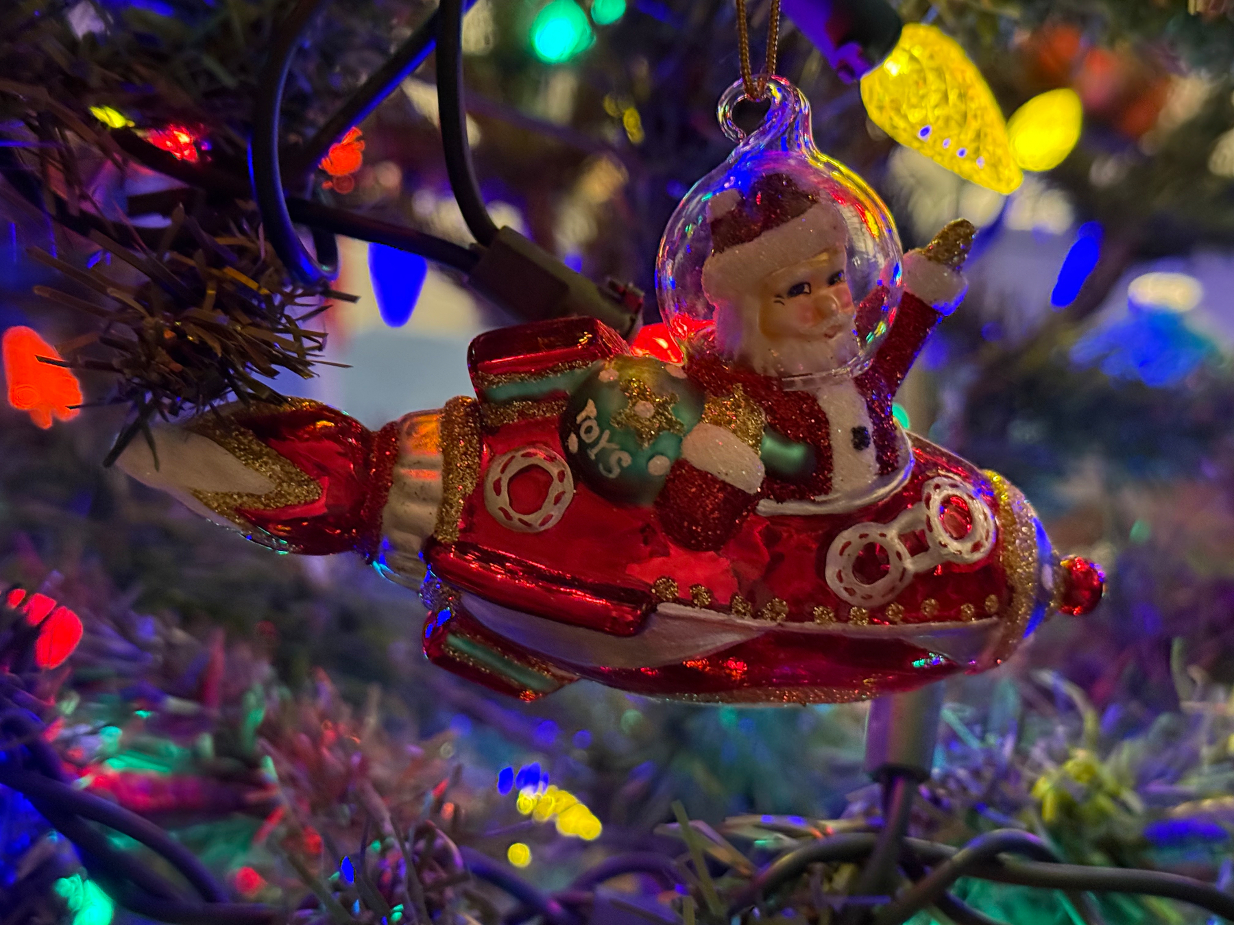 A festive ornament depicting Santa Claus in a spaceship is hanging on a Christmas tree decorated with colorful lights.