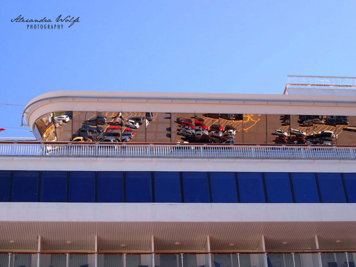 A cruise ship features a reflective glass facade that mirrors several vehicles parked below.