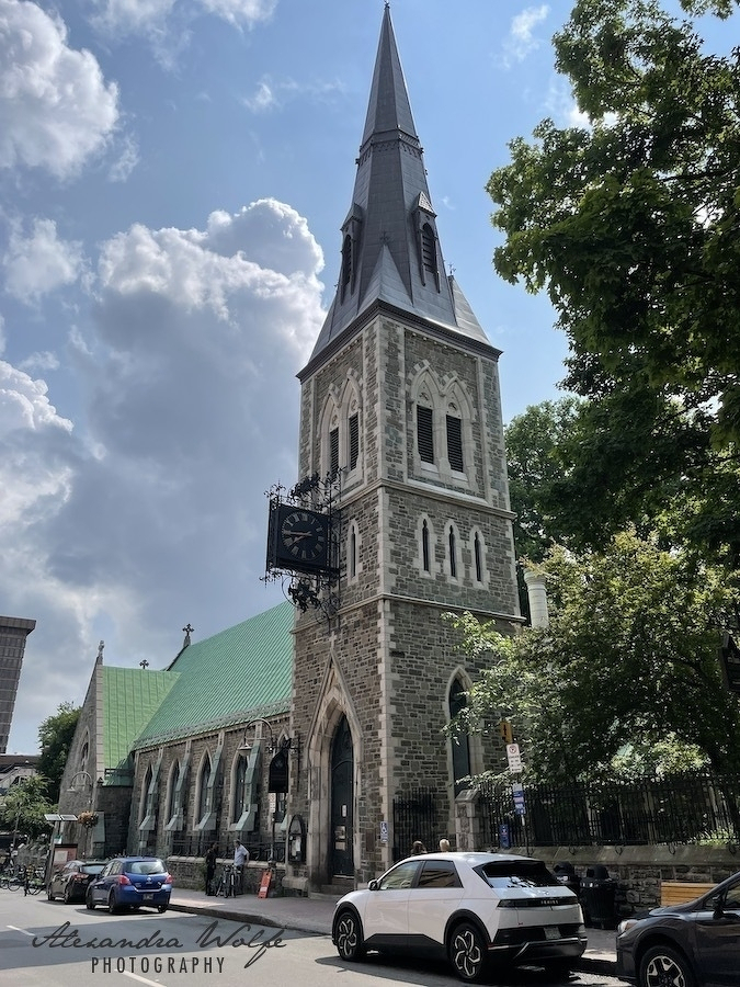 A historic stone church with a tall spire and an ornate clock on its tower is situated along a street lined with parked cars and trees.