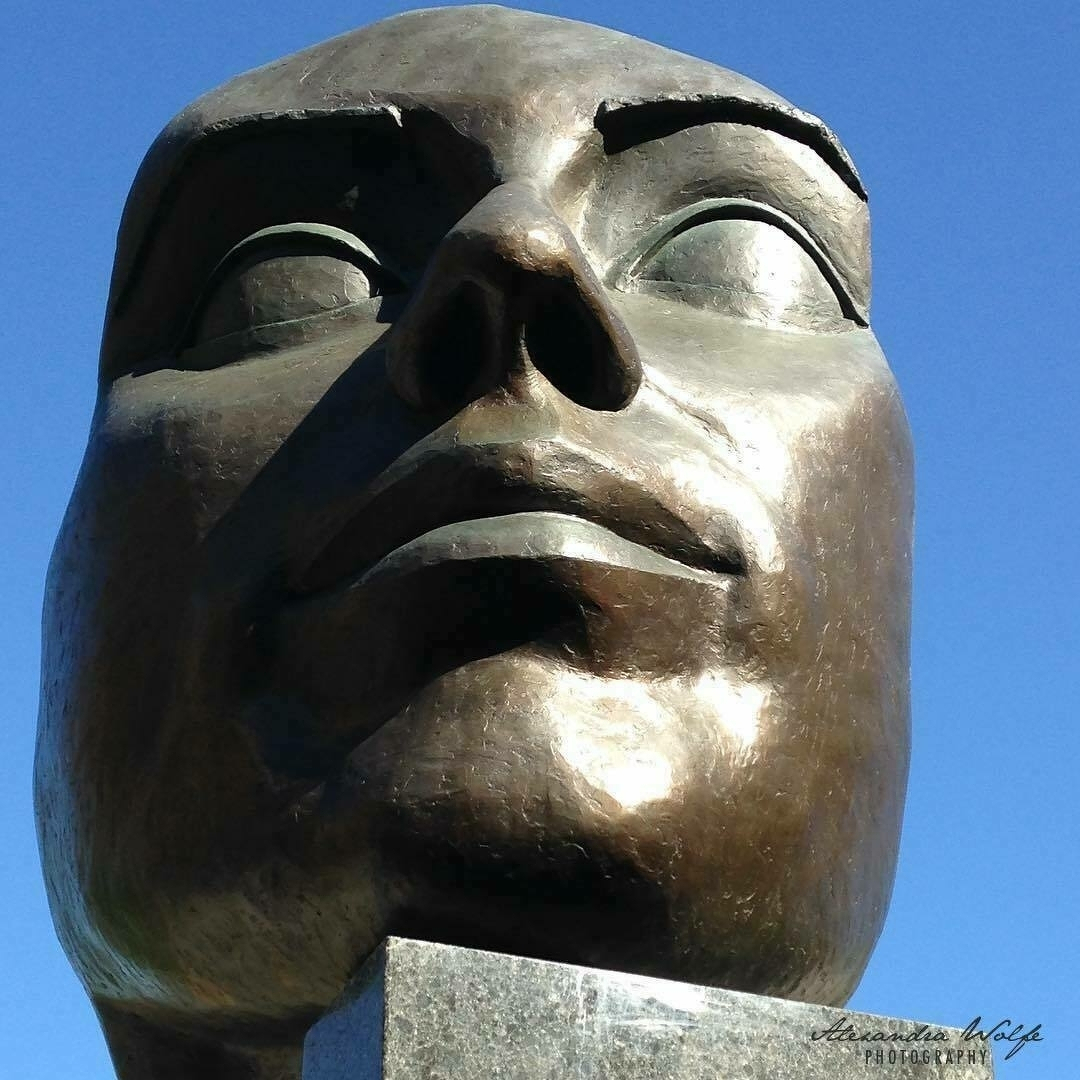 A large bronze sculpture of a human face is shown against a clear blue sky.