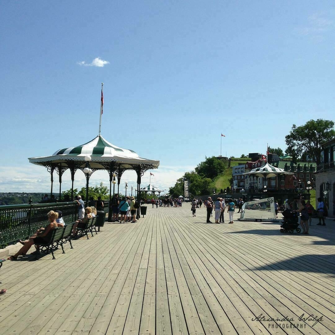 A sunny boardwalk with people walking and sitting on benches next to decorative pavilions and historic buildings.