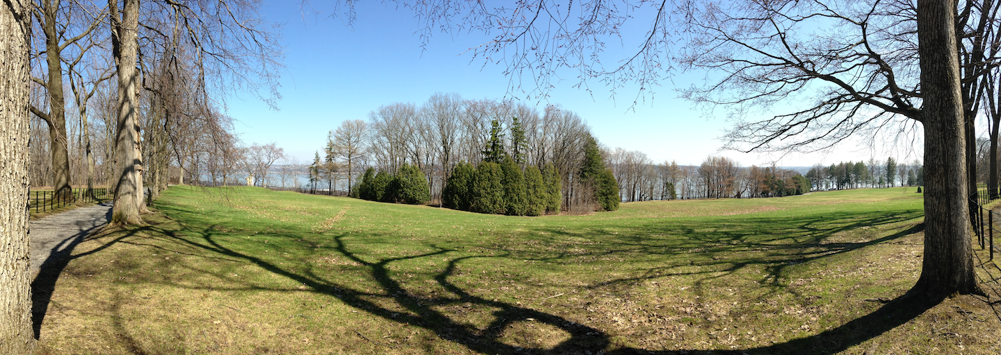 A vast grassy field is bordered by leafless trees and overlooks a distant body of water under a clear blue sky.