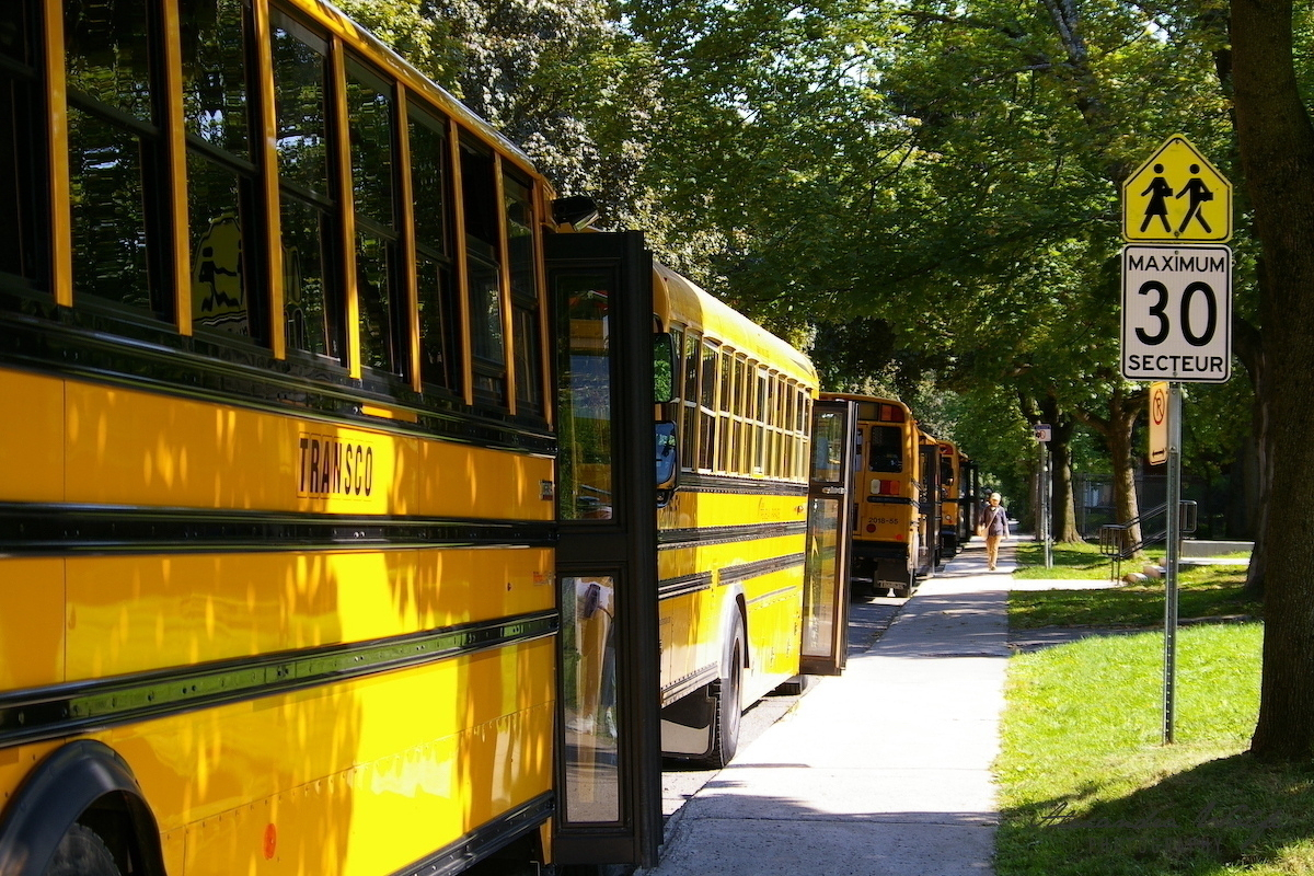 Several yellow school buses are parked along a tree-lined street next to a speed limit sign indicating a maximum of 30 km/h in a school zone.