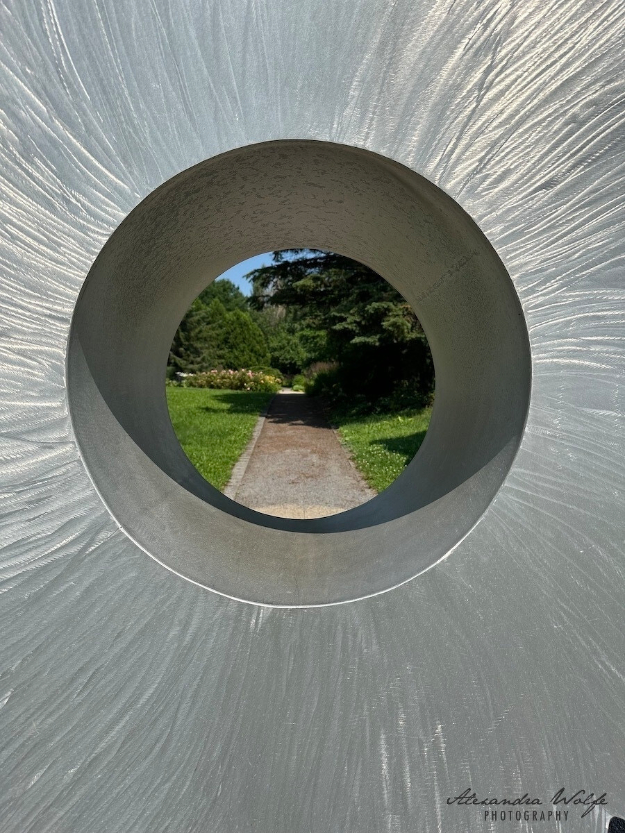 A circular stone sculpture frames a pathway surrounded by greenery and trees in a park setting.
