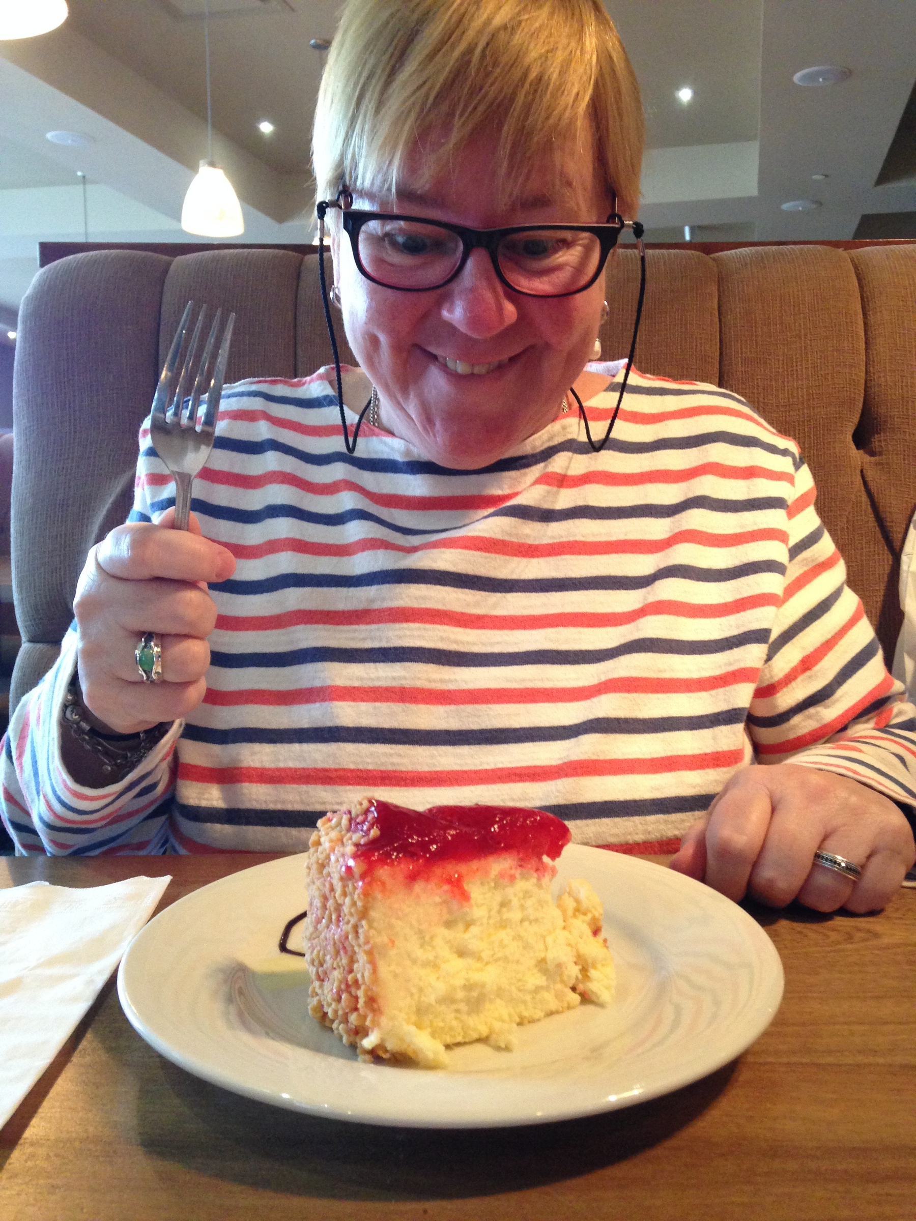 A person wearing glasses and a striped top is eagerly holding a fork and looking at a plate of cheesecake with strawberry topping.