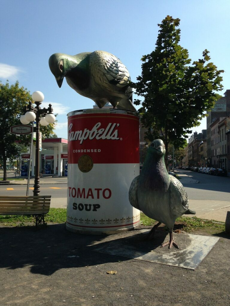 Two large pigeon sculptures are positioned on and near an oversized Campbell's Tomato Soup can in an outdoor park setting.