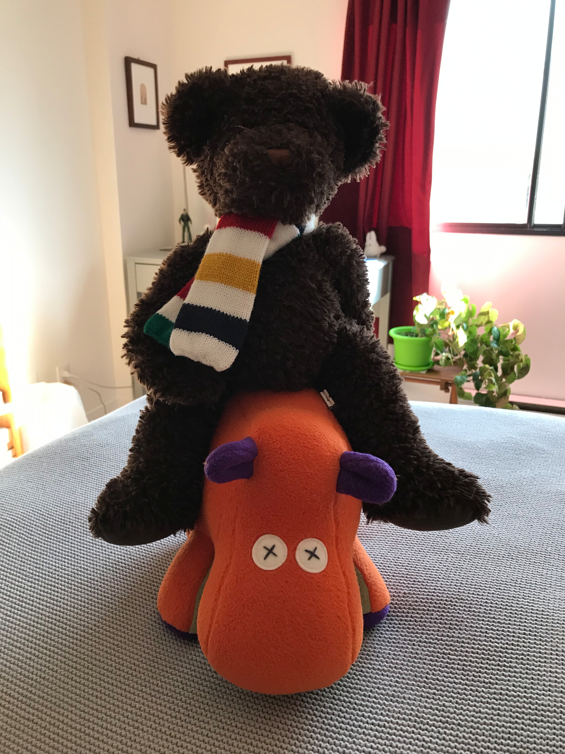 A brown teddy bear wearing a colorful scarf is sitting on an orange stuffed animal with purple details in a room with a plant and window.