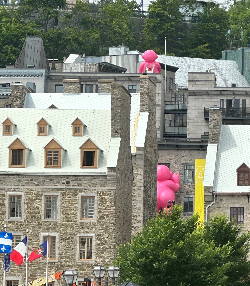 A large pink inflatable man is partially visible behind historic stone buildings with flags and trees in the foreground.