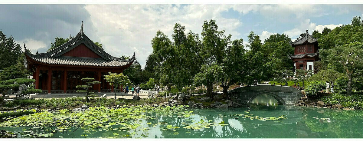 A tranquil garden scene features traditional Asian architecture, a pond with lily pads, a stone bridge, and lush greenery under a partly cloudy sky.