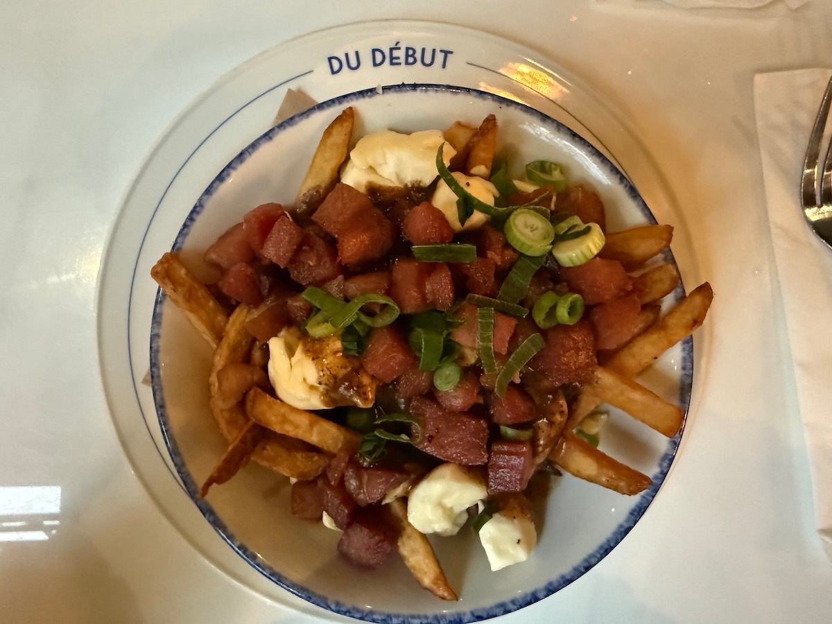 A plate of poutine topped with diced Gaspor ham, green onions, and cheese curds is served on a white plate labeled Du Début.