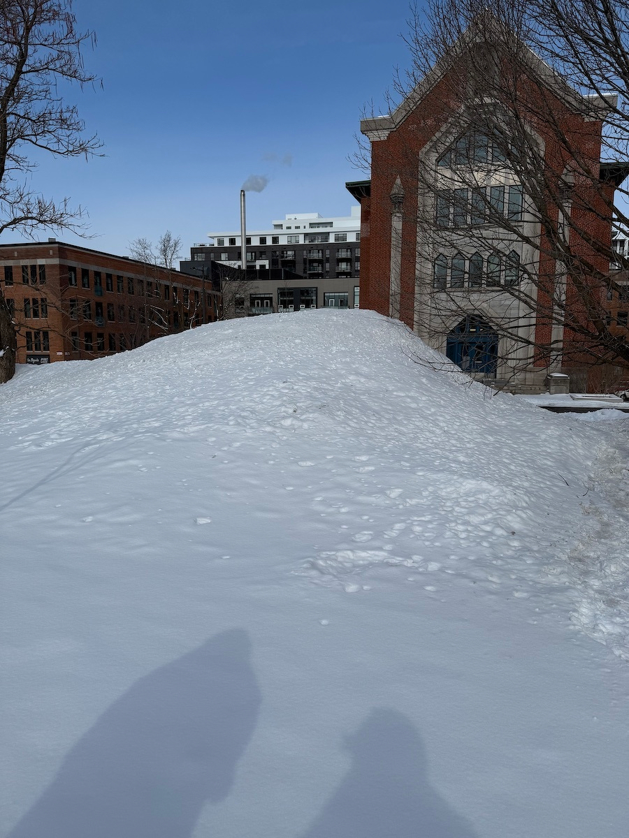 A snowy hill leads up to a large brick building with a pointed roof and adjacent industrial-style structures under a clear blue sky.
