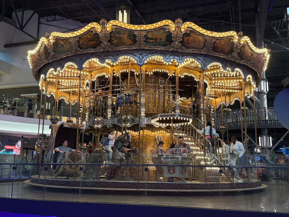 A beautifully illuminated, vintage-style double-decker carousel is situated indoors with people enjoying the ride.
