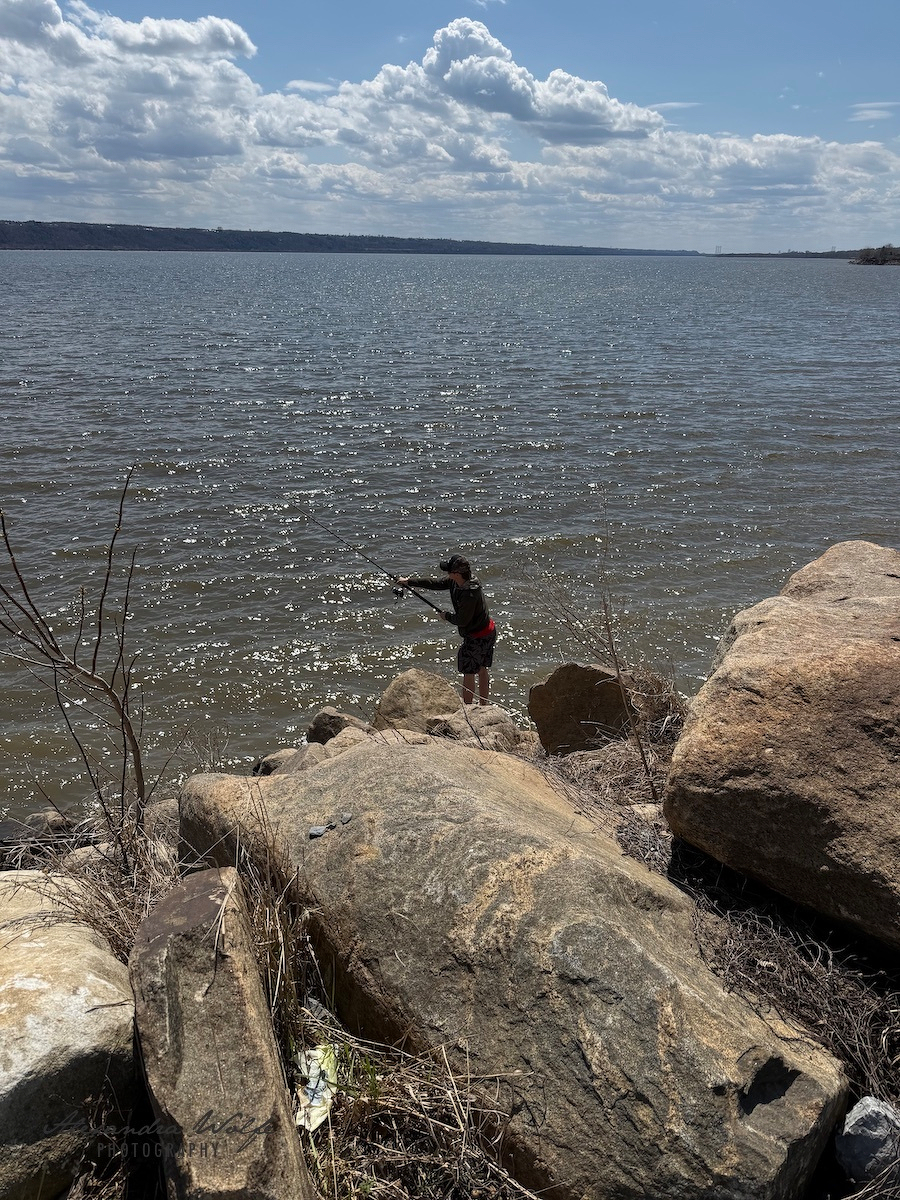 A person is fishing while standing on large rocks by the edge of a body of water under a partly cloudy sky.