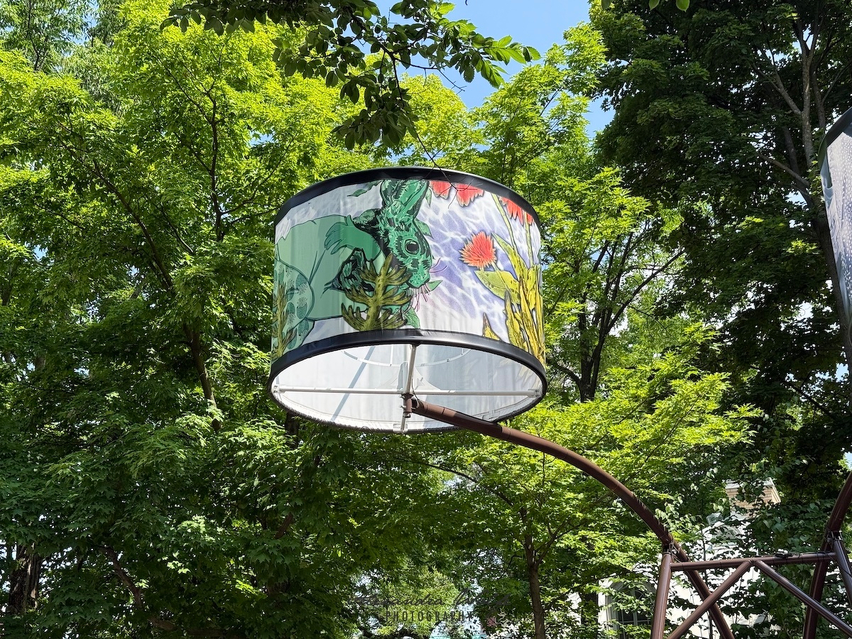 A large painted lampshade features a unique design like a large drum with colourful artwork depicting plants animals set against a backdrop of lush green trees.