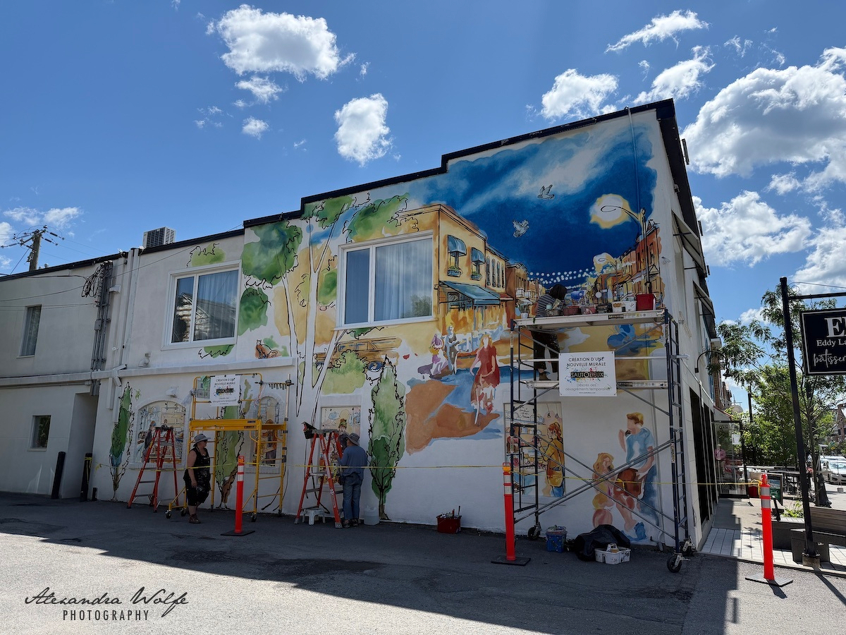 A colorful mural depicting a lively street scene with people, buildings, and a bright sky is being painted on the side of a building by several artists.