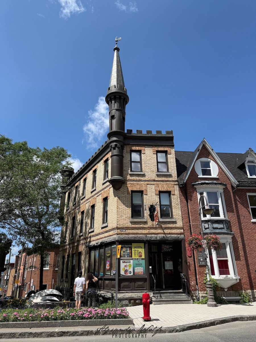 A unique three-story brick building with a tall, pointed turret stands on a street corner under a clear blue sky.