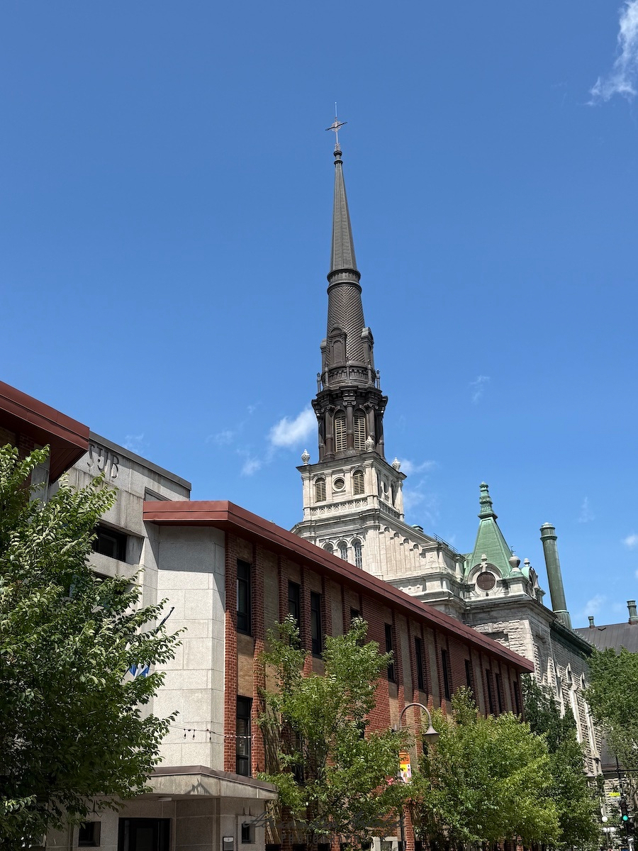 A tall, ornate church steeple rises above a combination of modern and historic buildings under a clear blue sky.