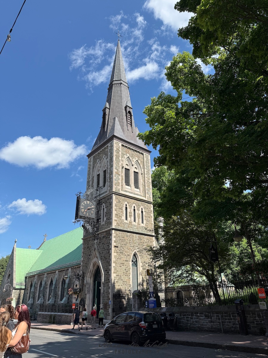 A tall stone church with a pointed steeple stands beside a street under a partly cloudy blue sky, with people walking and cars parked nearby.