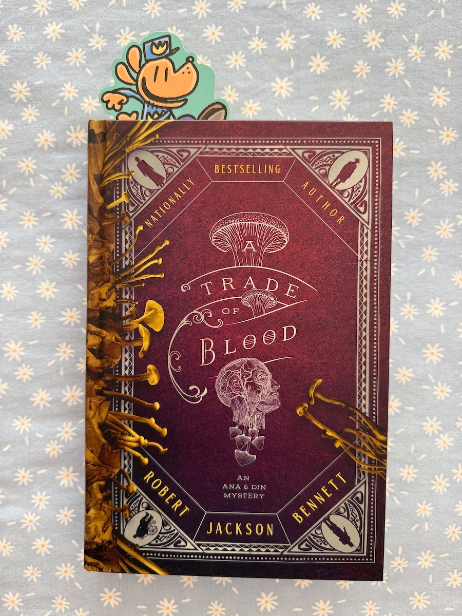 A hardcover book titled A Trade of Blood: An Ana & Din Mystery by Robert Jackson Bennett is shown with a decorative cover featuring mushroom illustrations and a skull design.