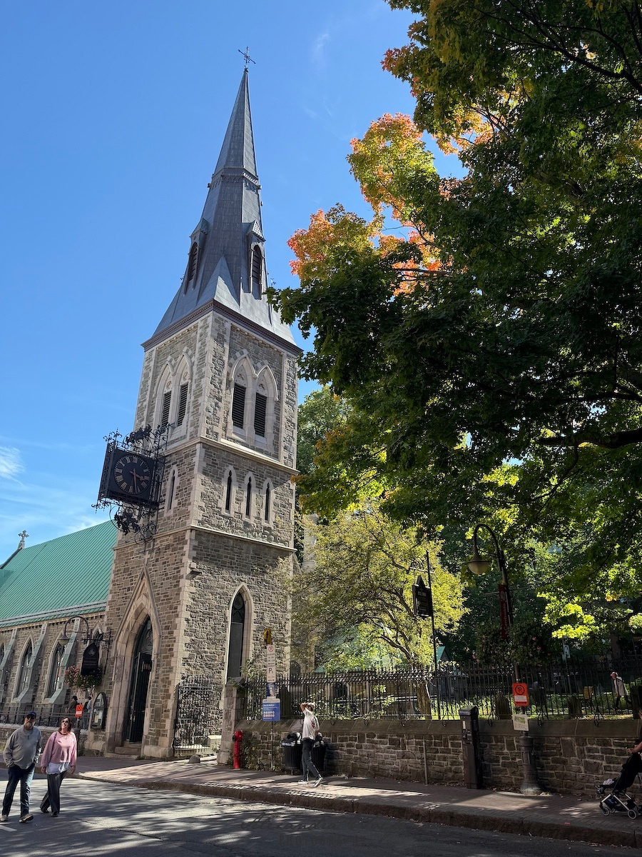 A historic stone church with a tall steeple and clock stands beside a leafy street where people are walking on a sunny day.