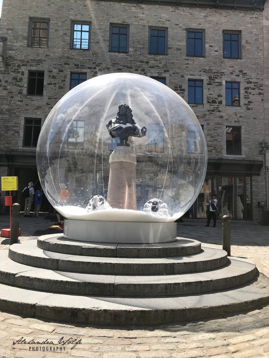 A sculpture of Louis XIV is encased in a large transparent snowglobe on a stone pedestal in a cobblestone courtyard.