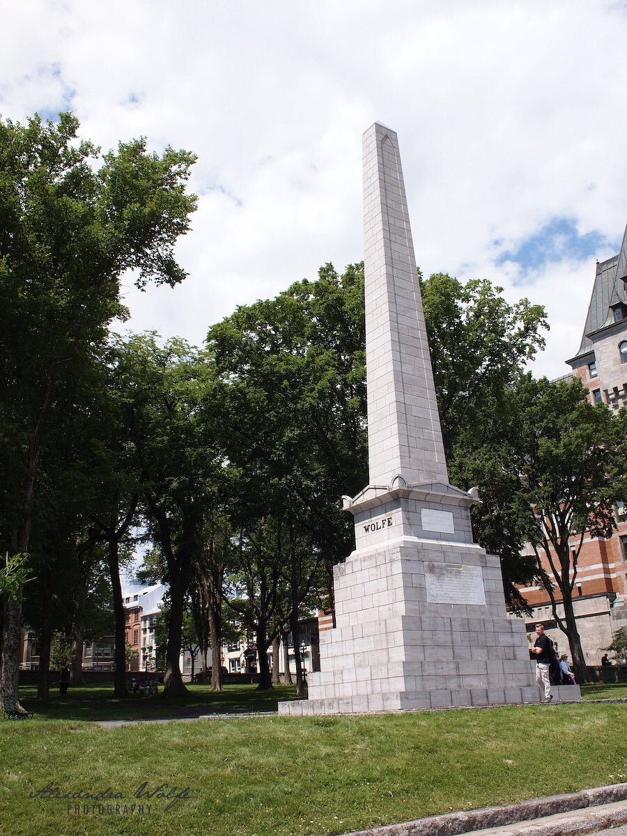 A tall, white stone obelisk stands on a grassy area surrounded by trees and buildings under a partly cloudy sky.