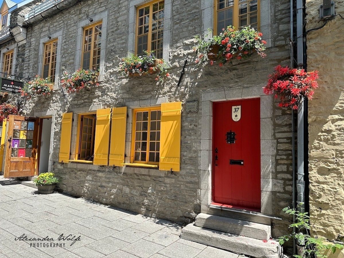 A charming stone building features yellow window shutters, vibrant red flower boxes, and a bright red door numbered 37, situated on a sunlit street.
