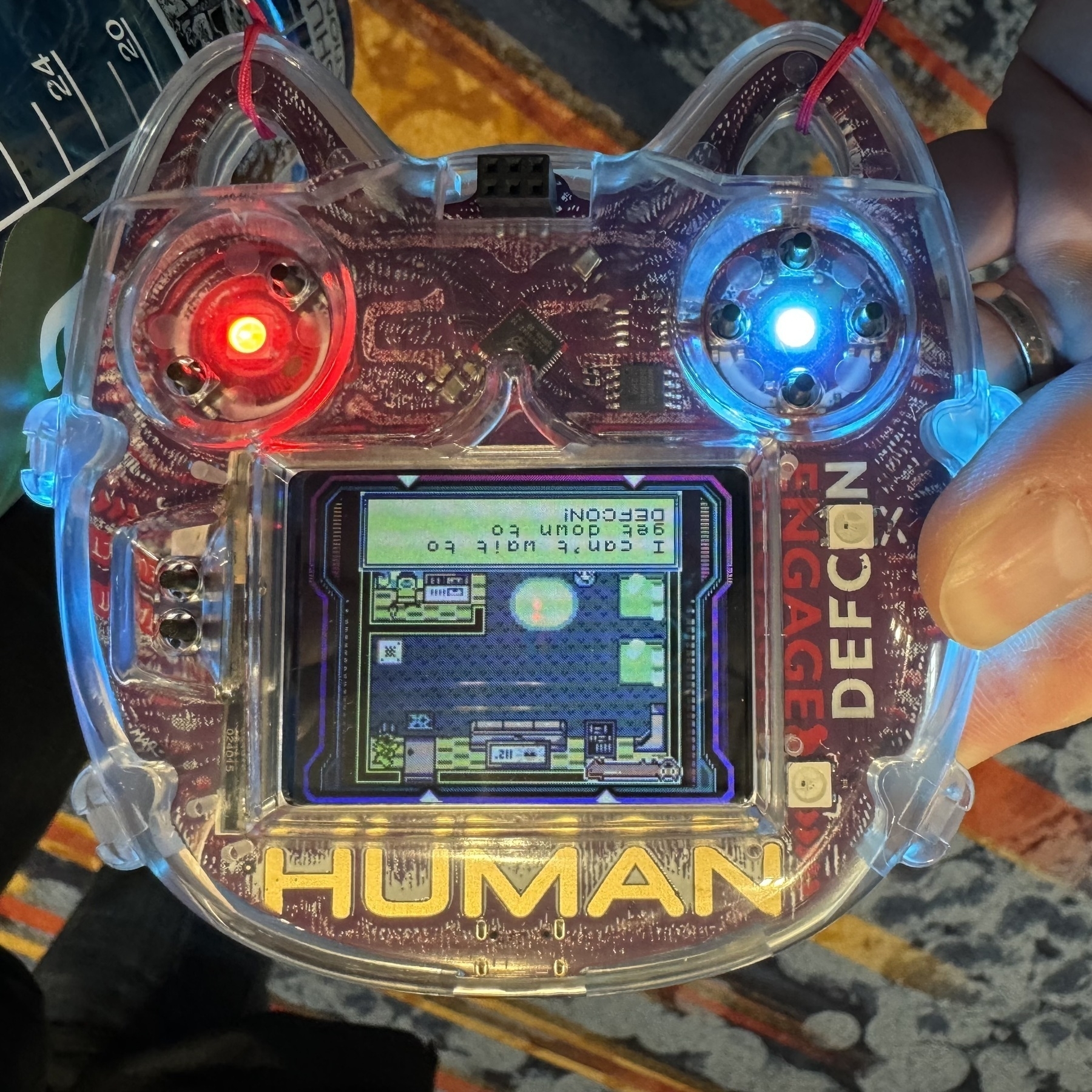 A roughly cat-shaped clear badge with electronics inside, displaying a Gameboy-style game. It’s glowing with bluish LEDs, except for the red left eye.