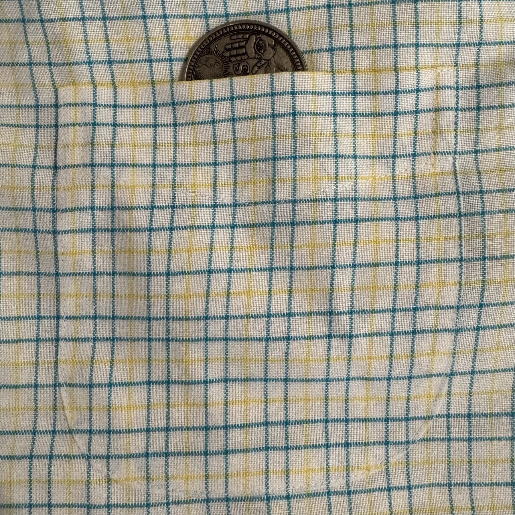 A photo of a shirt pocket. The cloth is white with a grid of small blue and yellow lines. The pocket is so aligned with the shirt that it’s nearly invisible, except that a large coin is sticking out the top to highlight where pocket ends and shirt begins. Someone was paying attention at that factory.