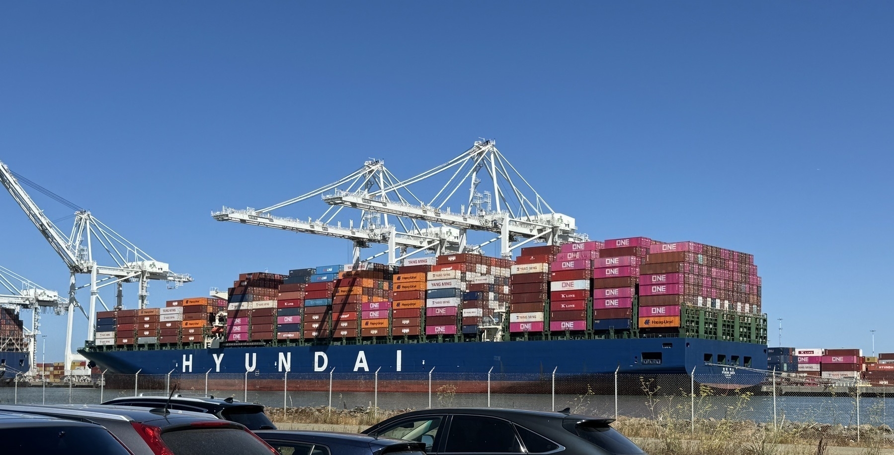 A giant Hyundai container ship loaded with pink and orange and brown and other brightly colored containers in front of some giant cranes and a clear blue sky. It sits across a channel and some car roofs are in the foreground.