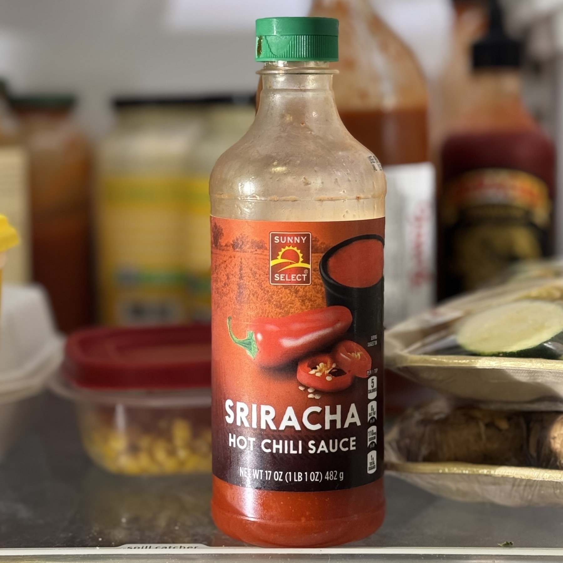 An open refrigerator with a half-empty bottle of sriracha on the very front of the middle shelf, label facing forward saying “SRIRACHA”.&10;&10;There is another open bottle of it in the background. It is not the 3rd one that the kid just now opened.