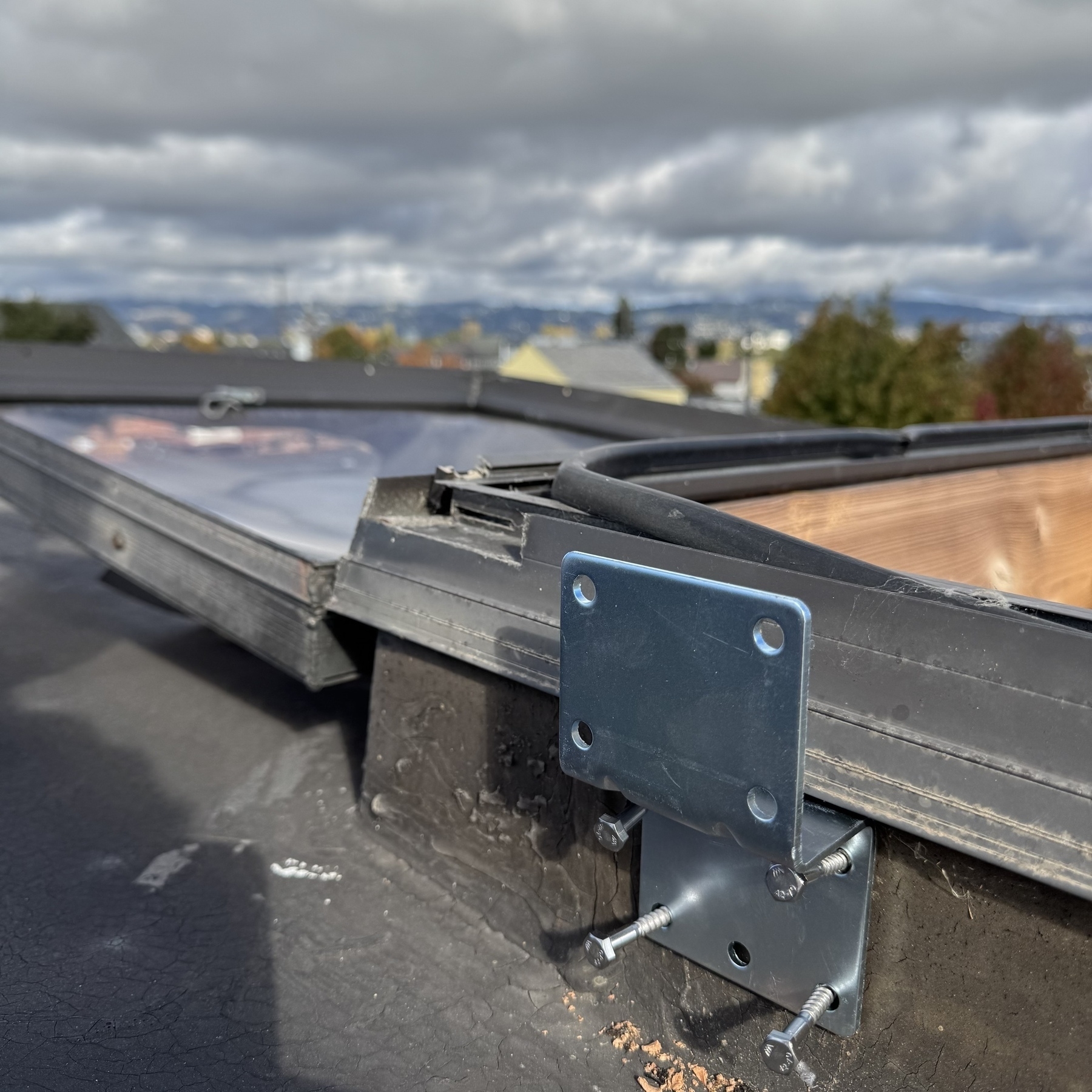 A silver bracket mount attached to the edge of a skylight on top of a roof. The screws are still sticking out because it hasn’t been bolted in yet. A cloudy sky hovers over the distant Oakland hills skyline.