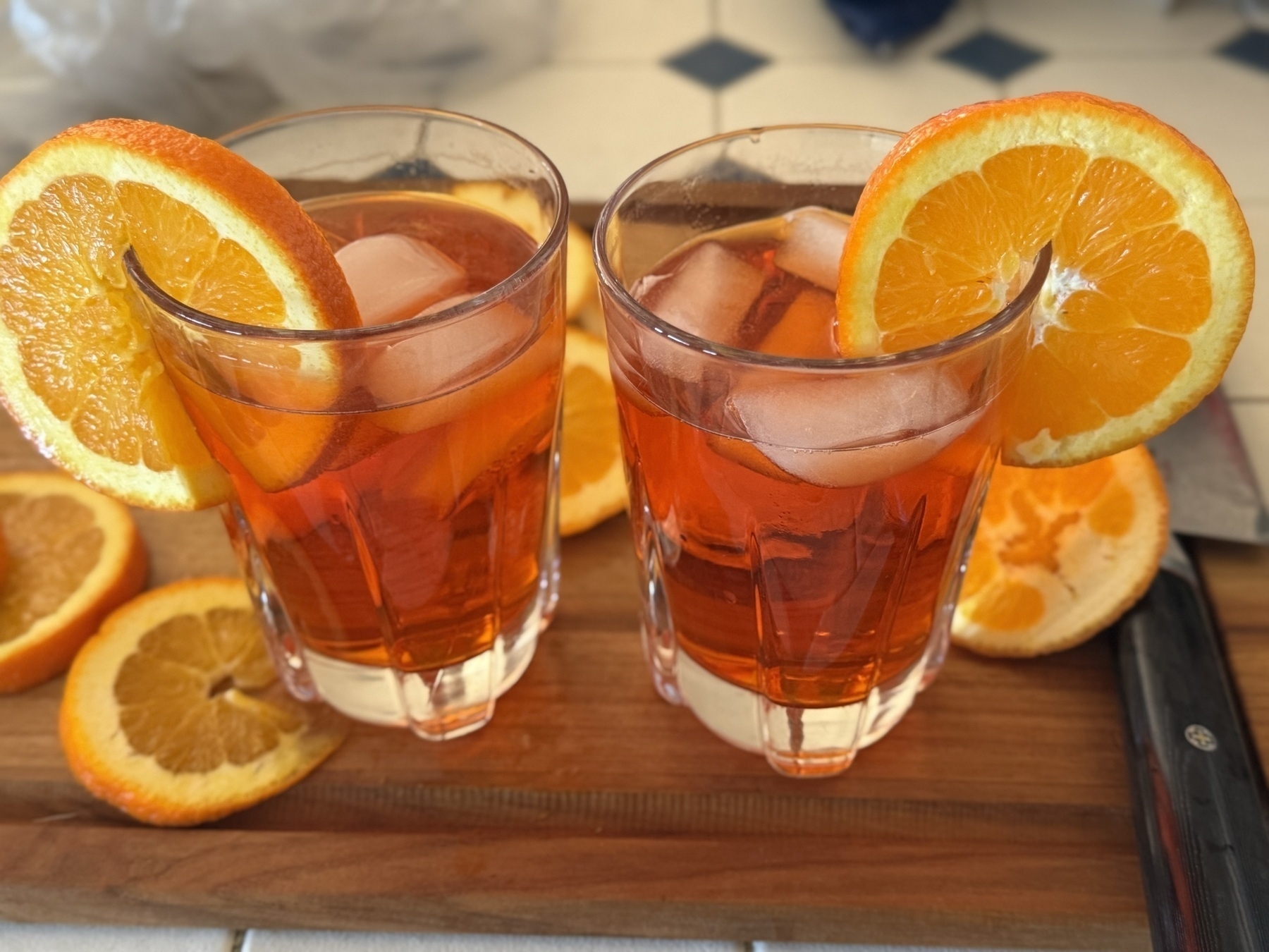 Two glasses are filled with dark orange liquid and topped with ice and orange slices. They’re sitting on a cutting board surrounded by more orange slices.