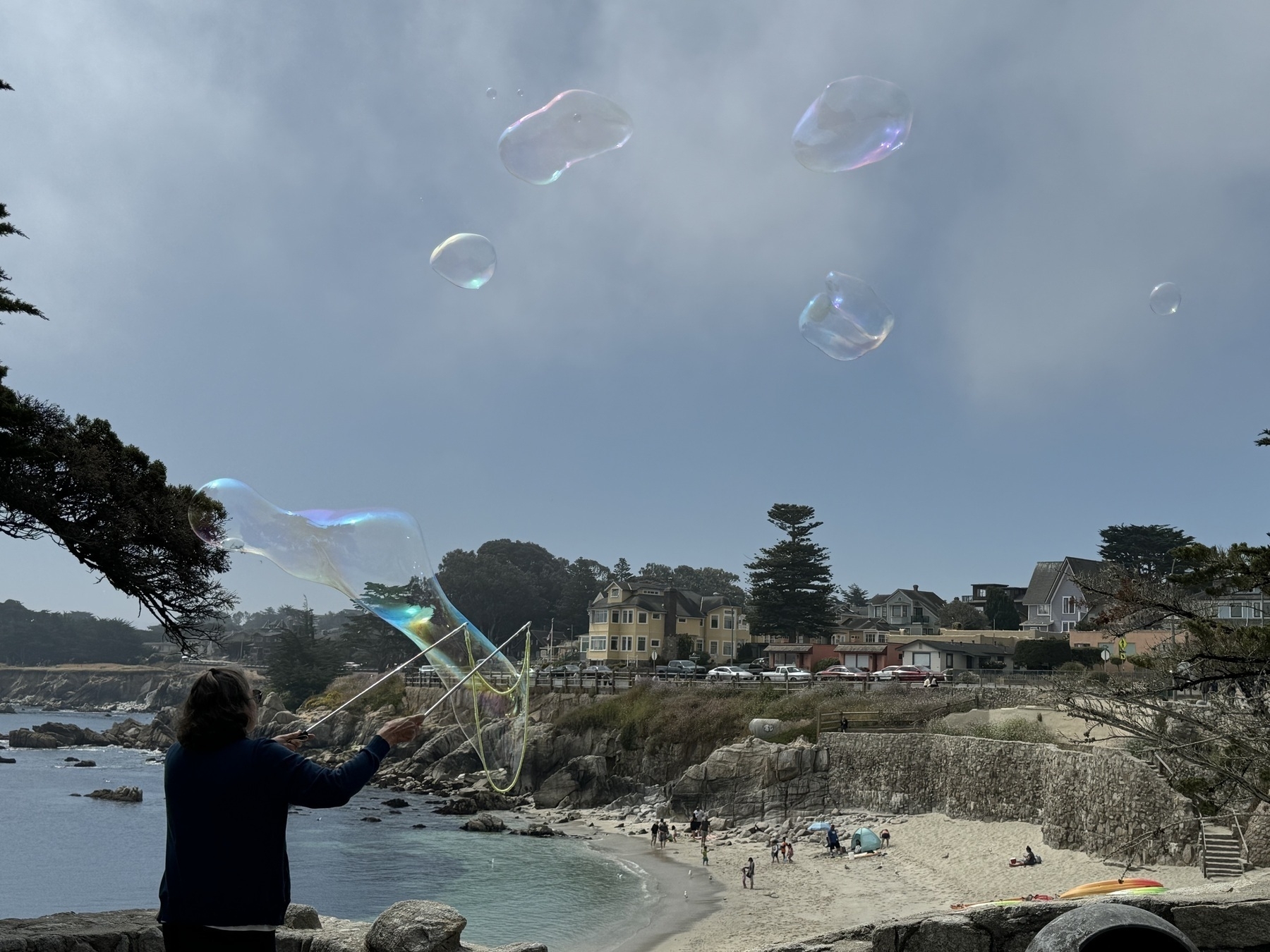 A sandy stretch of beach in Pacific Grove, CA. In the foreground someone is blowing enormous bubbles, like several feet across, with a bucket of soap and a loop of rope. The giant bubbles are shimmery and reflective.