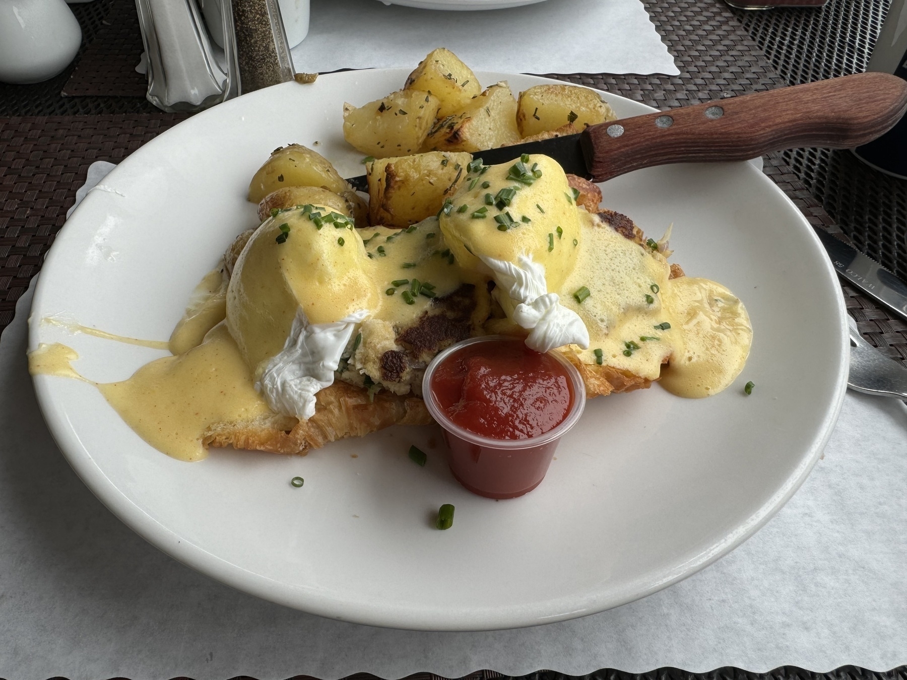 A plate of poached eggs Benedict over a croissant, and rosemary roasted potatoes.