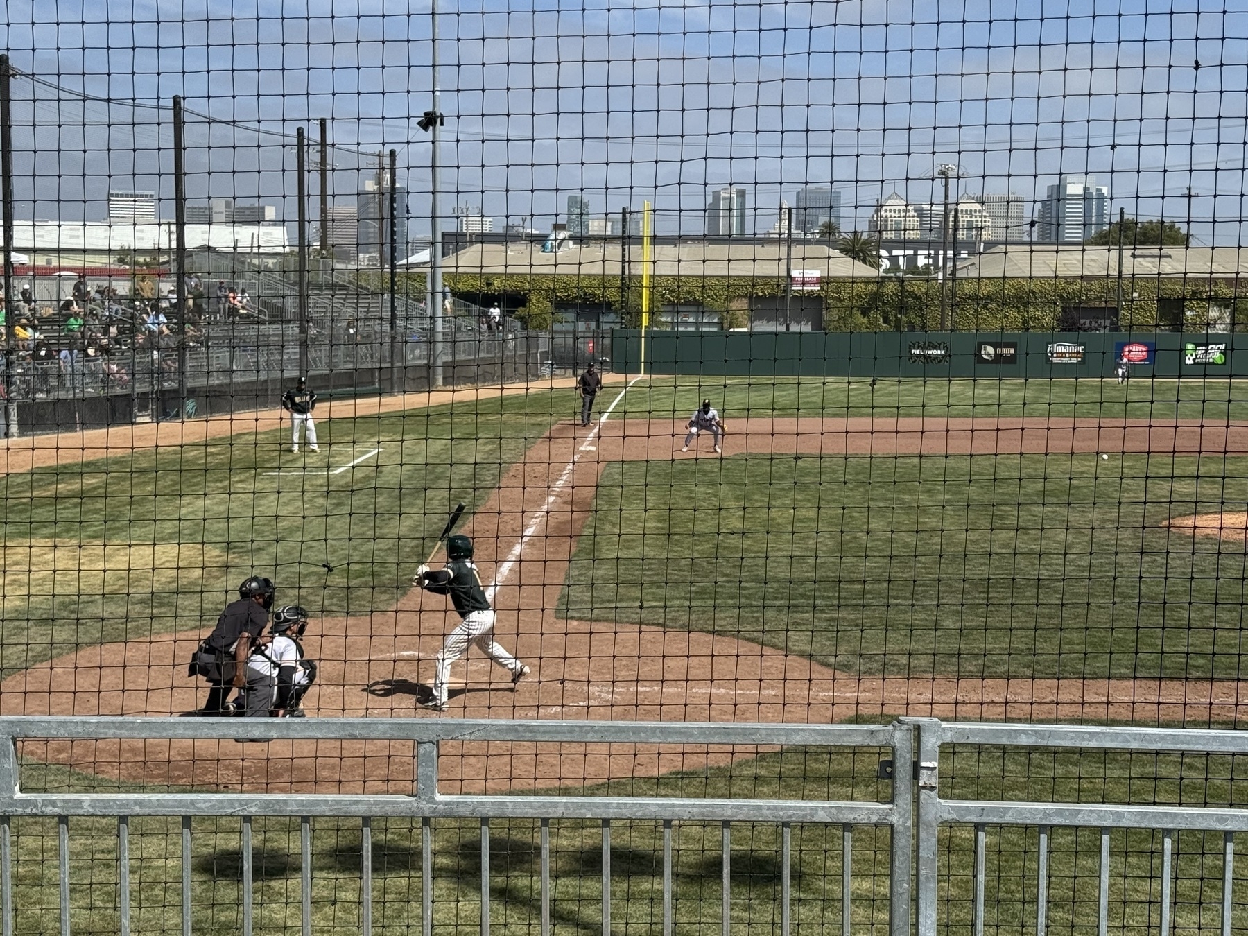 A pro baseball game from behind home plate, looking down the 3rd base line. Downtown Oakland is in the background.